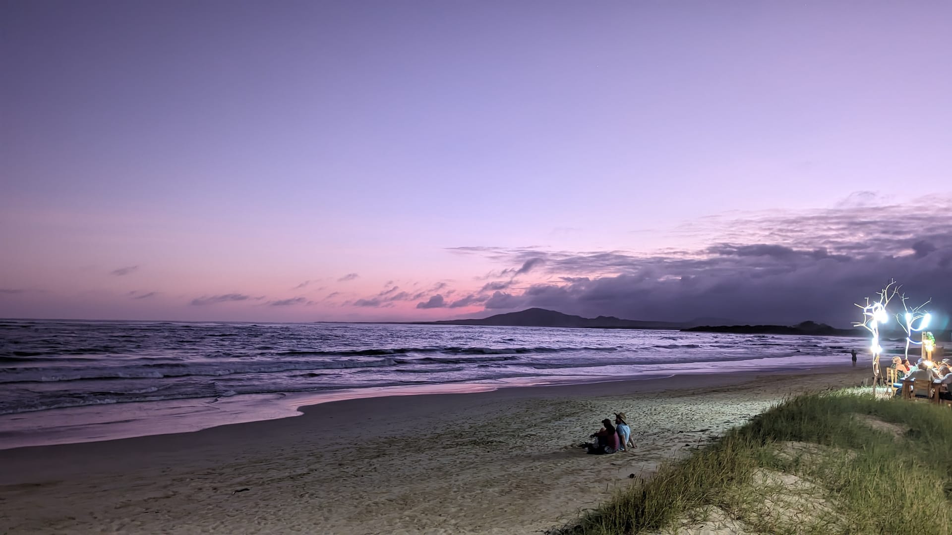 Lavender and pink dusk over the beach at Puerto Villamil, Isabela, with a couple sitting on the sand and beach-bar lights glowing