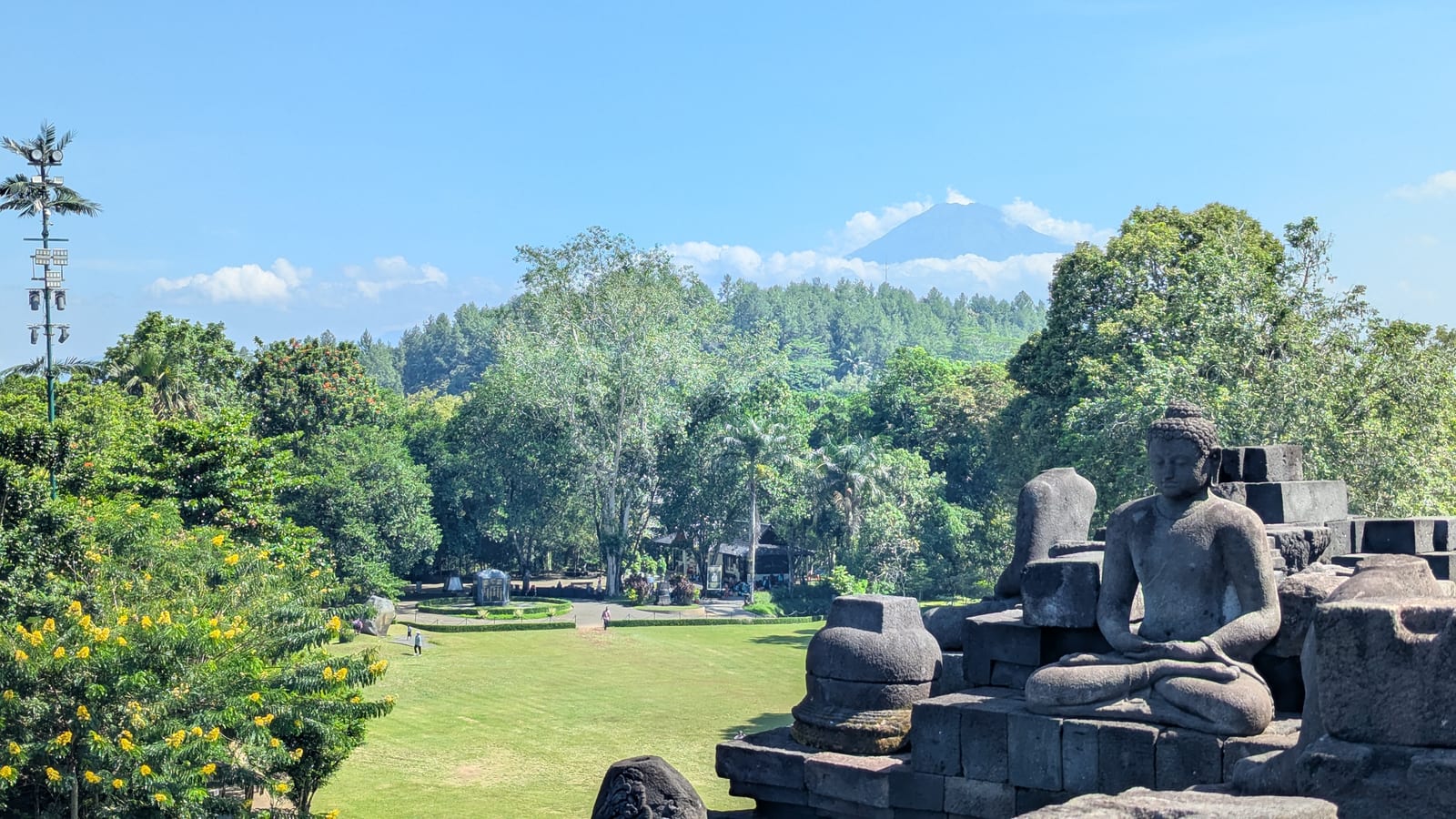 Wide view of Borobudur temple with stupas, lawn, and mountains under blue sky